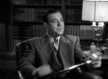 Movie still from “The Wolf Man” (1941), directed by George Waggner – A black and white photo of a man sitting in a chair in front of bookshelves; Medium shot, Low angle