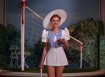 Movie still from “The Women” (1939), directed by George Cukor – A woman wearing a hat and holding a tennis racquet in front of a volleyball net; Medium shot, Low angle