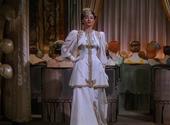 Movie still from “The Women” (1939), directed by George Cukor – A woman in a white dress standing in front of a table; Medium shot, Low angle