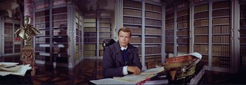 Movie still from “The Wonderful World of the Brothers Grimm” (1962), directed by Henry Levin – A man sitting at a desk in a library; Medium shot, Low angle