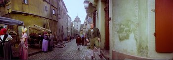 Movie still from “The Wonderful World of the Brothers Grimm” (1962), directed by Henry Levin – A man in a green jacket is standing in the street; Wide shot, Low angle