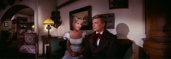 Movie still from “The Wonderful World of the Brothers Grimm” (1962), directed by Henry Levin – A man and a woman sitting next to each other on a couch; Medium shot, Over the shoulder angle
