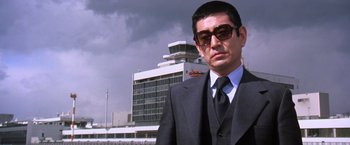Movie still from “The Yakuza” (1974), directed by Sydney Pollack – A man in a suit and tie standing in front of an airport; Medium shot, Low angle