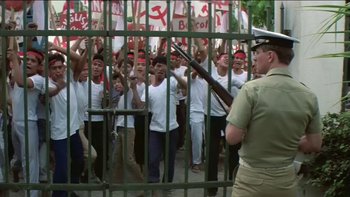 Movie still from “The Year of Living Dangerously” (1982), directed by Peter Weir – A group of people in front of a fence with a gun; Wide shot, High angle