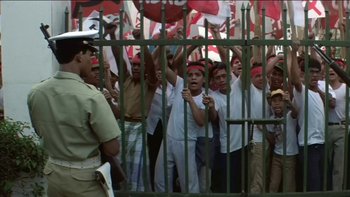 Movie still from “The Year of Living Dangerously” (1982), directed by Peter Weir – A group of people standing in front of a metal fence; Wide shot, High angle