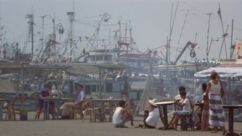 Movie still from “The Year of Living Dangerously” (1982), directed by Peter Weir – A group of people sitting at a table in front of boats in the water; Extreme Wide shot, High angle