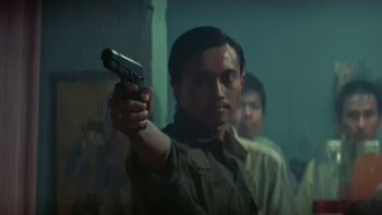 Movie still from “The Year of Living Dangerously” (1982), directed by Peter Weir – A man holding a gun in a room with other people; Medium shot, Low angle