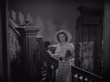 Movie still from “The Young in Heart” (1938), directed by Gilbert Pratt – A woman in a dress and a hat standing on a staircase; Medium shot, High angle