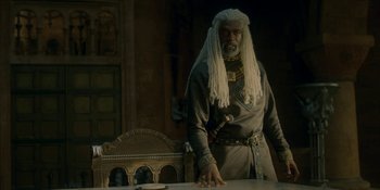 Movie still from “House of the Dragon” (2022), created by George R.R. Martin – An old man with long white hair standing in front of a table; Medium shot, Low angle