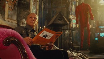 Movie still from “The Zero Theorem” (2013), directed by Terry Gilliam – A man sitting in a chair reading a book; Medium shot, Low angle