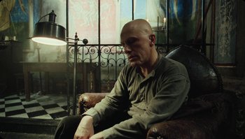 Movie still from “The Zero Theorem” (2013), directed by Terry Gilliam – A bald man sitting in a chair in a room; Medium shot, Low angle