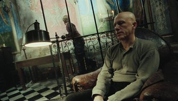 Movie still from “The Zero Theorem” (2013), directed by Terry Gilliam – A man sitting on a chair in front of a metal fence; Medium shot, Low angle