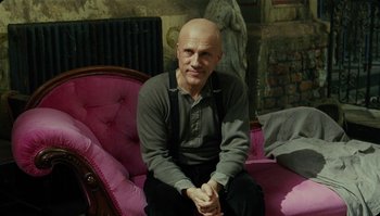 Movie still from “The Zero Theorem” (2013), directed by Terry Gilliam – A bald man sitting on a pink couch; Medium shot, Low angle