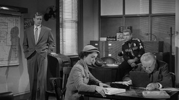 Movie still from “Them!” (1954), directed by Gordon Douglas – A woman sitting at a table with papers in front of two men; Wide shot, Over the shoulder angle
