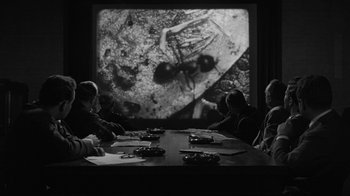 Movie still from “Them!” (1954), directed by Gordon Douglas – A group of people sitting at a table watching a television screen; Wide shot, High angle