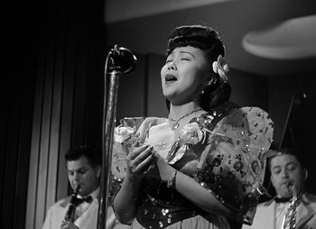 Movie still from “They Were Expendable” (1945), directed by Robert Montgomery – A woman is singing into a microphone while a man watches; Medium shot, Low angle
