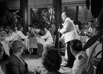 Movie still from “They Were Expendable” (1945), directed by Robert Montgomery – A man standing on a stage in front of a group of people; Wide shot, Over the shoulder angle
