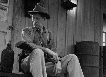 Movie still from “They Were Expendable” (1945), directed by Robert Montgomery – An old man sitting on a bench wearing a hat and holding a gun; Medium shot, Low angle
