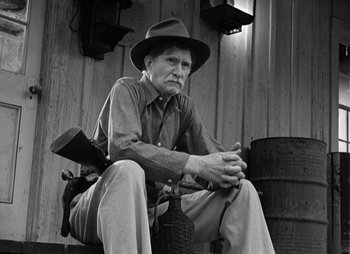 Movie still from “They Were Expendable” (1945), directed by Robert Montgomery – An older man sitting on a barrel wearing a hat; Medium shot, Low angle