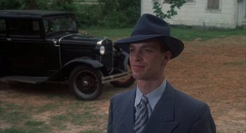 Movie still from “Thieves Like Us” (1974), directed by Robert Altman – A man in a suit and hat standing in front of an old car; Medium shot, Low angle