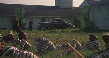 Movie still from “Thieves Like Us” (1974), directed by Robert Altman – An old car parked in the grass next to a building; Wide shot, High angle