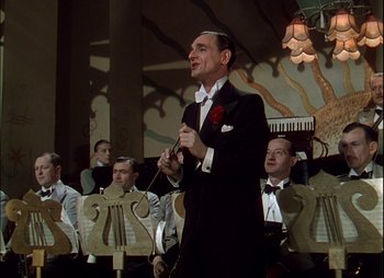 Movie still from “This Happy Breed” (1944), directed by David Lean – A man in a suit and bow tie holding a stick in front of a group of people; Medium shot, Low angle