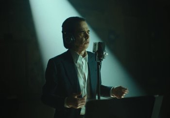 Movie still from “This Much I Know to Be True” (2022), directed by Andrew Dominik – A man in a suit and headphones is speaking into a microphone; Medium shot, Low angle