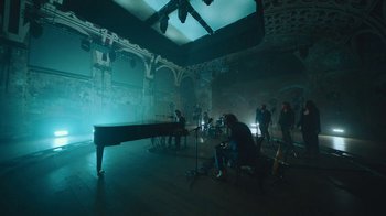Movie still from “This Much I Know to Be True” (2022), directed by Andrew Dominik – A group of people standing around a grand piano in a room; Extreme Wide shot, High angle
