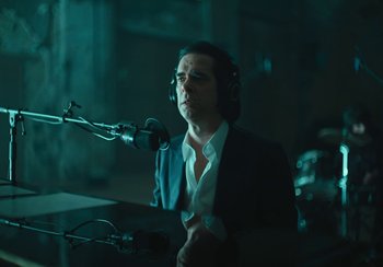 Movie still from “This Much I Know to Be True” (2022), directed by Andrew Dominik – A man in a suit and headphones is sitting in front of a microphone; Medium shot, Over the shoulder angle