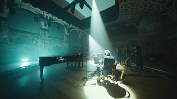 Movie still from “This Much I Know to Be True” (2022), directed by Andrew Dominik – A group of people in a room with a piano; Extreme Wide shot, High angle