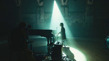 Movie still from “This Much I Know to Be True” (2022), directed by Andrew Dominik – A person sitting at a piano in a dark room; Wide shot, Low angle