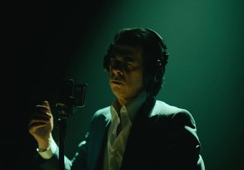 Movie still from “This Much I Know to Be True” (2022), directed by Andrew Dominik – A man in a suit is holding a microphone and wearing headphones; Medium shot, Low angle