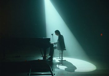 Movie still from “This Much I Know to Be True” (2022), directed by Andrew Dominik – A man sitting at a piano in a room with a light shining on him; Wide shot, Low angle