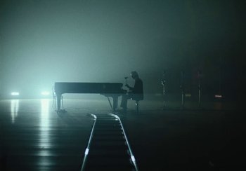 Movie still from “This Much I Know to Be True” (2022), directed by Andrew Dominik – A person sitting at a piano in a dark room; Extreme Wide shot, High angle