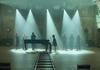 Movie still from “This Much I Know to Be True” (2022), directed by Andrew Dominik – A group of people standing around a grand piano; Extreme Wide shot, High angle