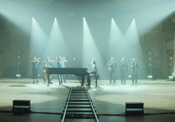Movie still from “This Much I Know to Be True” (2022), directed by Andrew Dominik – A group of people standing on a stage with a piano; Extreme Wide shot, High angle