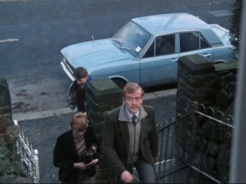 Movie still from “Threads” (1984), directed by Mick Jackson – A man and a woman sitting on steps next to a blue car; Wide shot, High angle