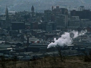 Movie still from “Threads” (1984), directed by Mick Jackson – Smoke billows out of a chimney on a city skyline; Extreme Wide shot, High angle