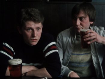 Movie still from “Threads” (1984), directed by Mick Jackson – Two men sitting at a table with a drink; Medium shot, Over the shoulder angle