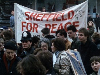 Movie still from “Threads” (1984), directed by Mick Jackson – A group of people gathered around a banner; Medium shot, High angle