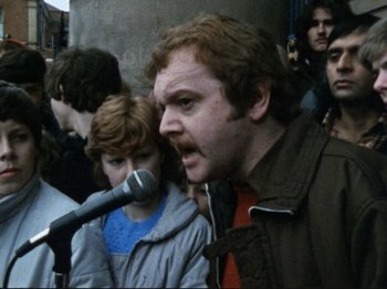 Movie still from “Threads” (1984), directed by Mick Jackson – A man with a microphone in front of a group of people; Close Up shot, High angle