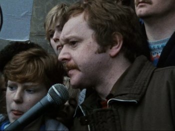 Movie still from “Threads” (1984), directed by Mick Jackson – A man with a beard is speaking into a microphone; Close Up shot, Over the shoulder angle