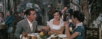 Movie still from “Three Coins in the Fountain” (1954), directed by Jean Negulesco – A group of people sitting at a table with a glass of wine; Medium shot, Over the shoulder angle