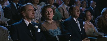 Movie still from “Three Coins in the Fountain” (1954), directed by Jean Negulesco – An older woman wearing a blue dress in front of a group of people; Medium shot, Over the shoulder angle