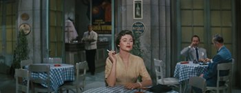 Movie still from “Three Coins in the Fountain” (1954), directed by Jean Negulesco – A woman sitting at a table with a cigarette in her mouth; Medium shot, Over the shoulder angle