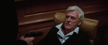Movie still from “Three Days of the Condor” (1975), directed by Sydney Pollack – An older man sitting in a chair holding a gun in his hand; Close Up shot, Over the shoulder angle