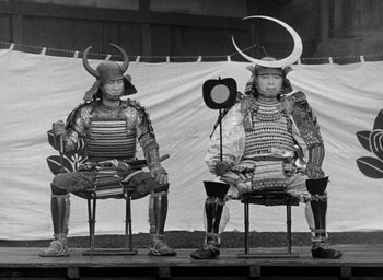 Movie still from “Throne of Blood” (1957), directed by Akira Kurosawa – A couple of men sitting on top of chairs wearing armor; Wide shot, High angle