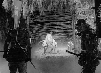Movie still from “Throne of Blood” (1957), directed by Akira Kurosawa – A man sitting in a straw hut surrounded by two other men; Wide shot, High angle