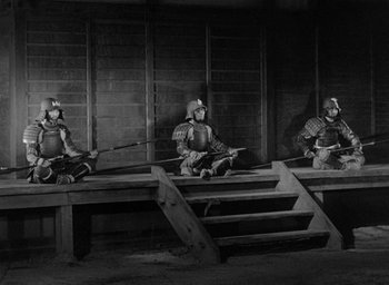 Movie still from “Throne of Blood” (1957), directed by Akira Kurosawa – A group of men sitting next to each other on some wooden steps; Wide shot, High angle