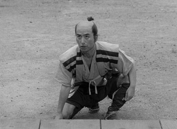Movie still from “Throne of Blood” (1957), directed by Akira Kurosawa – A man kneeling down on the ground with a bun in his hair; Medium shot, High angle